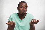 Photo of hesitant African American male with puzzled expression, shrugs shoulders in bewilderment, can`t find answer to difficult question, looks desperately at camera, isolated over white wall