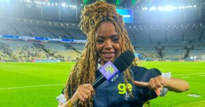 Jornalista negra Jordana Araújo sorri segurando microfone em estúdio de TV.